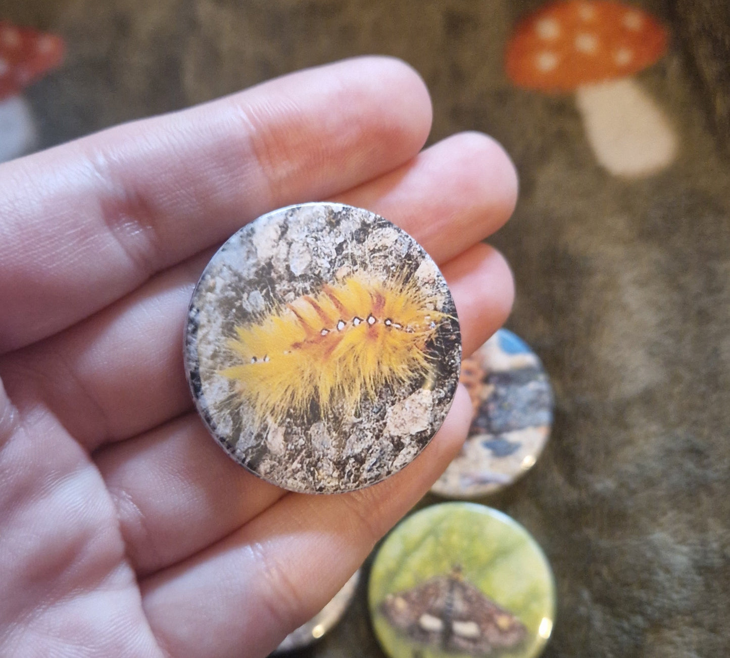 Close-up of a hand holding a round, pin badge with a colorful caterpillar design against a textured background.